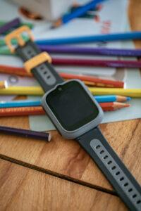 Graue TCL MOVETIME Kindersmartwatch mit schwarzem Display auf einem Holztisch, umgeben von bunten Buntstiften.