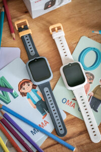 Zwei TCL MOVETIME Kindersmartwatches in Grau und Weiß liegen auf Kinderbüchern neben bunten Stiften.